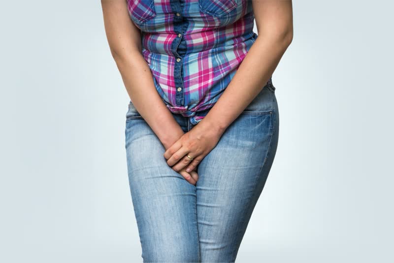 Donna con jeans e camicia a quadretti rosa che si tiene le mani all'altezza del pube ad emulare un'urgenza di minzione per simboleggiare l'incontinenza urinaria